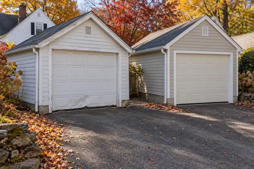 Older white garage door with a dented bottom section and sagging seal beside a newer almond door