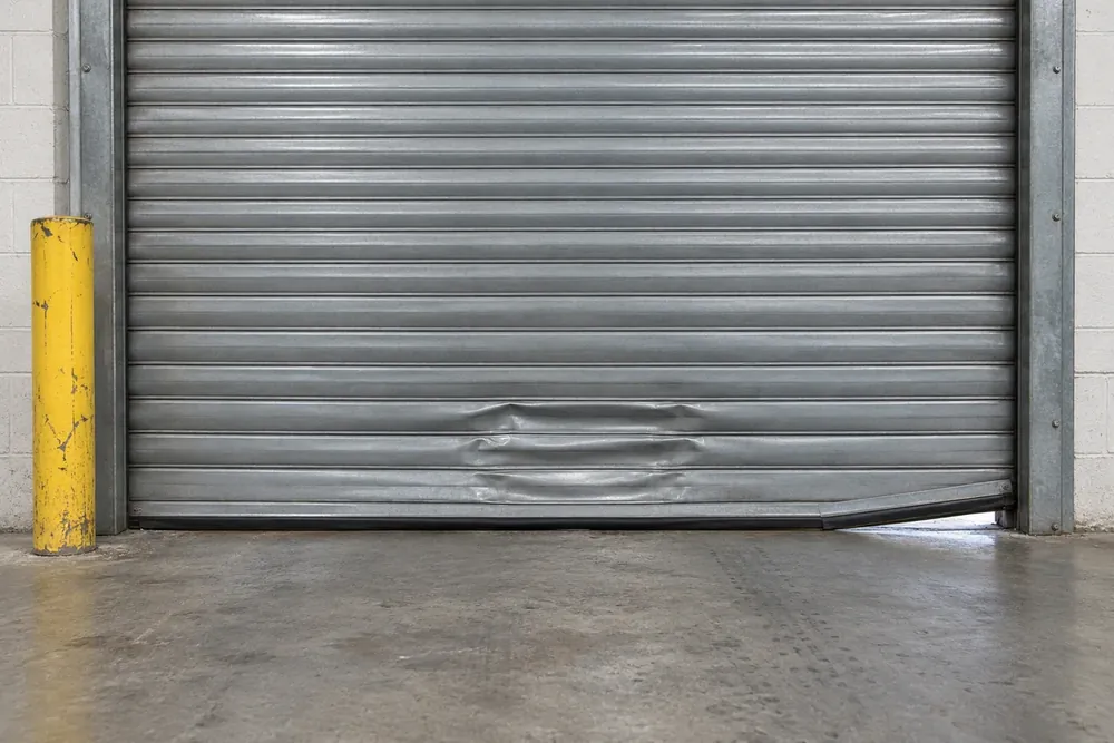 Dented lower slats and a bent bottom bar leave daylight beneath a warehouse rolling steel door