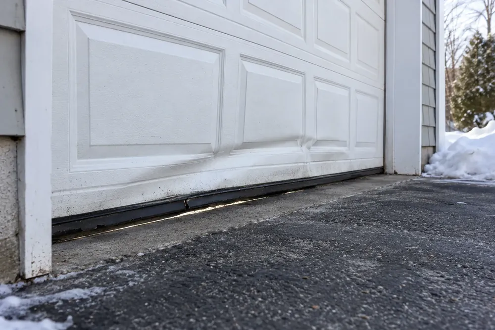 Dented white bottom section with worn rubber seal and daylight underneath, snow beside the driveway